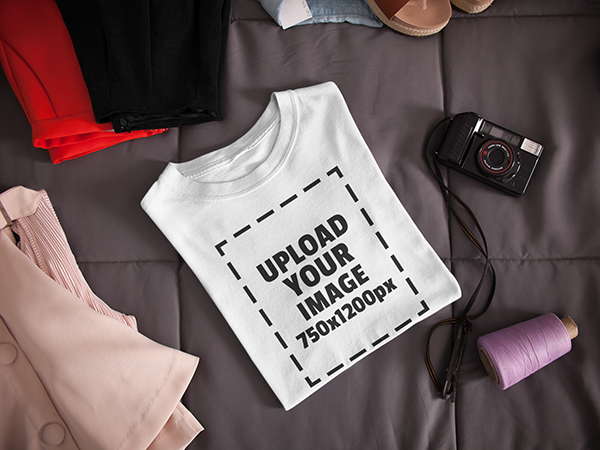 Folded t-shirt mockup lying next to a camera and clothes on a bed